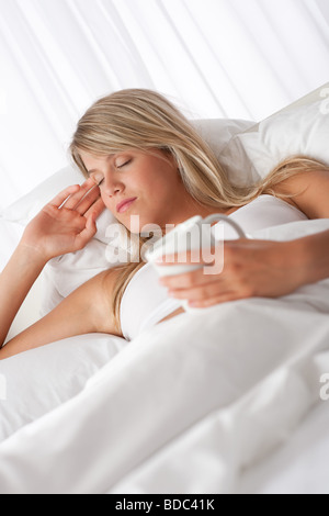 Blonde Frau im weißen Bett halten Tasse Kaffee mit geschlossenen Augen Stockfoto