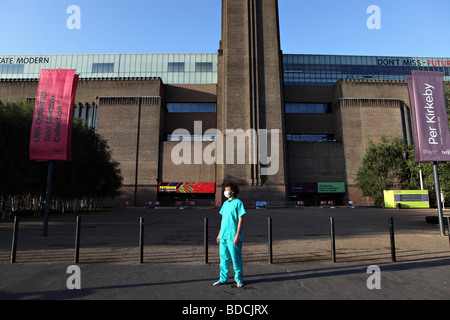 Ein junger Mann mit chirurgischer Mundschutz und medizinische einheitliche steht immer noch vor der Tate Modern in London. Stockfoto