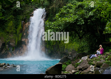 Weibliche Wanderer ruht unter einem Wasserfall entlang des pulsierenden blauen Rio Celeste Flusses im Vulkan-Nationalpark Tenorio, Costa Rica. Stockfoto