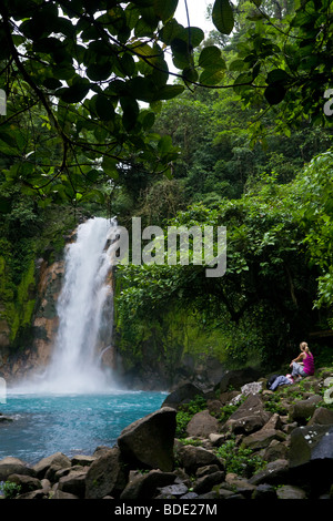 Weibliche Wanderer ruht unter einem Wasserfall entlang des pulsierenden blauen Rio Celeste Flusses im Vulkan-Nationalpark Tenorio, Costa Rica. Stockfoto