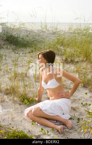 Junge Frau im weißen Bikini-Oberteil und Rock sitzt auf Sanddünen Strand Stockfoto