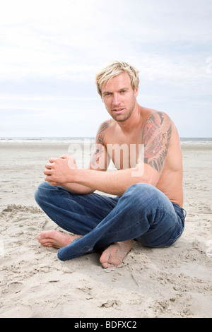 Junger Mann sitzt am Strand mit offenen Brustkorb Stockfoto