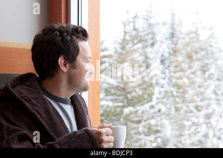 Mann im Bademantel hält eine Tasse Kaffee und blickte aus dem Fenster Stockfoto