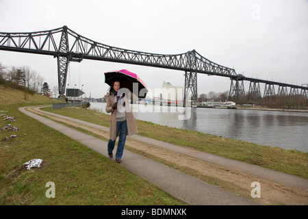 Junge Frau nass 30-35 Jahre, zu Fuß mit einem bunten Regenschirm entlang des Nord-Ostsee-Kanals an einem kalten Wintertag, Schleswig-Holstein, Stockfoto