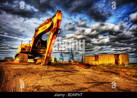 Große gelbe Bagger mit Caterpillar tracks auf der Baustelle mit Kränen in Ferne und bedrohlich dunkle Wolken unter blauem Himmel Stockfoto