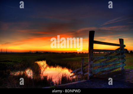 Sonnenuntergang über Elmley Sümpfe mit den Swale überqueren und die Kingsferry Brücke im Hintergrund und Reflexionen auf den Sonnenuntergang in einem Graben Stockfoto