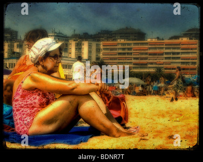 Eine alte Frau mit Baden Kostüm und weißer Hut sitzt an einem Strand mit Hotels im Hintergrund Stockfoto