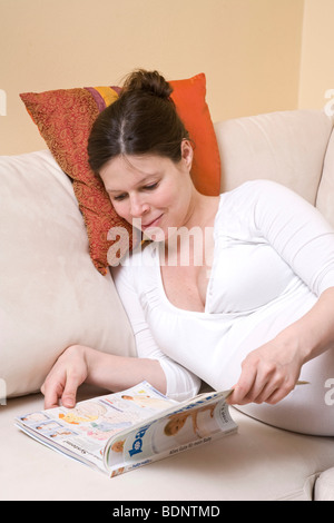 Schwangere Frau bequem auf dem Sofa liegend und ein Baby-Magazin lesen Stockfoto
