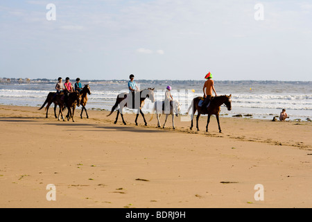 Eine Reitschule geht nach einer Kerbe am Strand und in der Brandung. Fahrer sind alle unterschiedlichen Alters und Könnens. Stockfoto