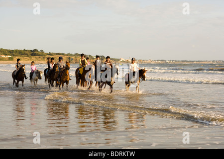 Eine Reitschule geht nach einer Kerbe am Strand und in der Brandung. Fahrer sind alle unterschiedlichen Alters und Könnens. Stockfoto