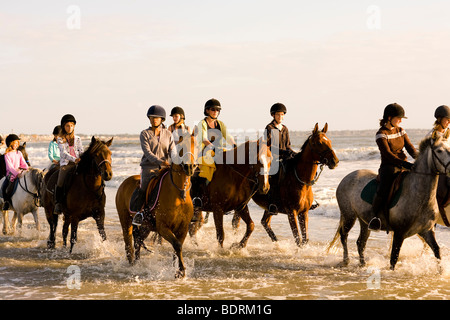 Eine Reitschule geht nach einer Kerbe am Strand und in der Brandung. Fahrer sind alle unterschiedlichen Alters und Könnens. Stockfoto