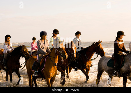 Eine Reitschule geht nach einer Kerbe am Strand und in der Brandung. Fahrer sind alle unterschiedlichen Alters und Könnens. Stockfoto