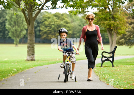 Horizontale nahe Porträt einer jungen Mutter hilft ihrem Sohn lernen, mit dem Fahrrad mit Stabilisatoren im park Stockfoto