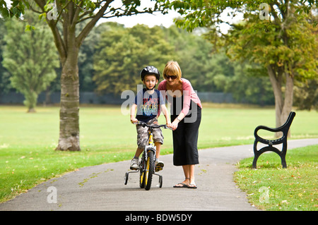 Horizontale nahe Porträt einer jungen Mutter hilft ihrem Sohn lernen, mit dem Fahrrad mit Stabilisatoren im park Stockfoto