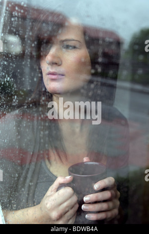 Dunkelhaarige Frau hält eine Kaffeetasse schauen einen Regen durchnässt Fenster. Stockfoto