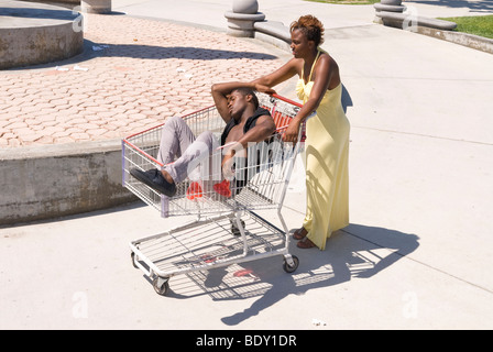 Eine Frau schiebt sie ohnmächtig Mann auf einem Gehweg in einem Einkaufswagen. Stockfoto