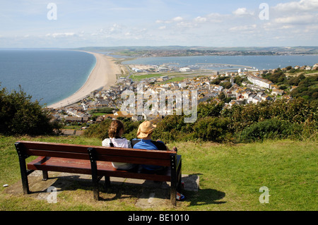 Paar sitzt auf einem Sitz auf der Isle of Portland Dorset England UK mit Blick auf Chesil Beach und Portland Harbour Stockfoto