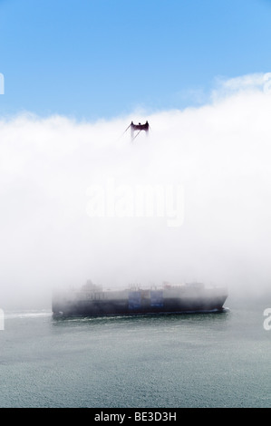 Golden Gate Bridge Tower in dichtem Nebel mit Frachtschiff San Francisco Kalifornien | SAN FRANCISCO, Kalifornien – Ein einzelner Turm der Golden Gate Bridge durchdringt ein Ufer aus dichtem Sommernebel, während ein Frachtschiff von Pascha Hawaii unter ihm in die Bucht von San Francisco gleitet. Die 1937 fertiggestellte Hängebrücke überspannt die Golden Gate Strait bis zum Marin County; die International Orange Towers ragen 746 Fuß (227 m) über dem Wasser. Dicker Meeresnebel, angetrieben von kaltem Auftrieb im Pazifik, bedeckt die Bucht regelmäßig während der Sommermonate. Stockfoto