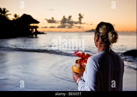 Eine Frau in einer weißen Tunika hat eine verzierte Kokosnuss in der Hand und blickt auf das Meer, Sunset Beach Resort in den Rücken, Mahe Isla Stockfoto