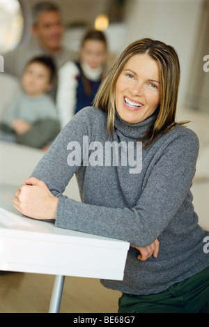 Frau sitzt am Boden lächelnd in die Kamera, Familie auf Sofa im Hintergrund Stockfoto