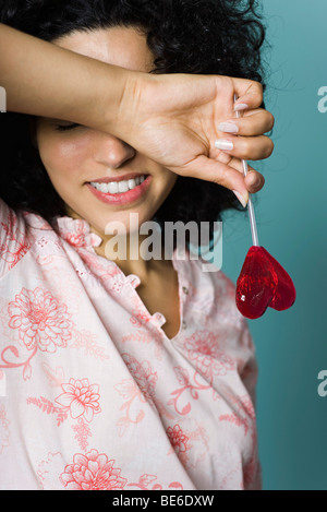 Frau Lächeln auf den Lippen, Augen mit Arm bedecken, hält herzförmige Lutscher Stockfoto