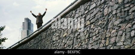 Geschäftsmann, stehend auf Wand mit erhobenen Armen, Hochhaus im Hintergrund Stockfoto