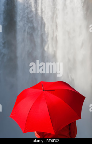 Eine Frau in einer roten Jacke gekleidet und tragen einen roten Regenschirm stehen vor einem Wasserfall. Stockfoto