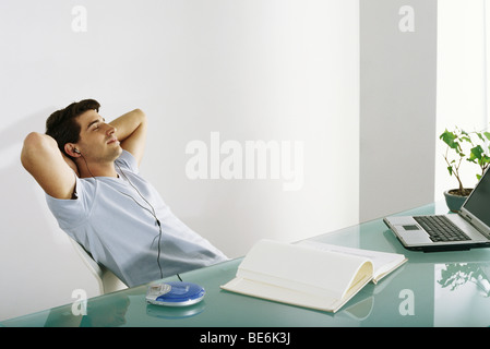 Mann am Schreibtisch gelehnt zurück mit Händen hinter dem Kopf, Kopfhörer anhören Stockfoto