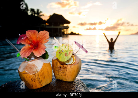 Zwei Kokosnüsse mit Cocktails und Dekorationen auf einem Granitfelsen bei Sonnenuntergang, in den Rücken, die, den eine Frau in das Meer, Seychellen steht Stockfoto