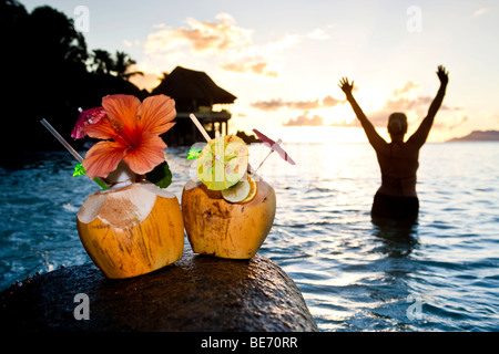 Zwei Kokosnüsse mit Cocktails und Dekorationen auf einem Granitfelsen bei Sonnenuntergang, in den Rücken, die, den eine Frau in das Meer, Seychellen steht Stockfoto