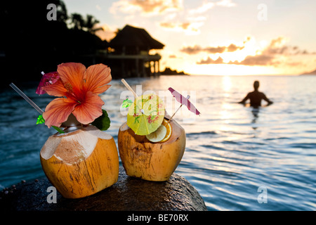 Zwei Kokosnüsse mit Cocktails und Dekorationen auf einem Granitfelsen bei Sonnenuntergang, in den Rücken, die, den eine Frau in das Meer, Seychellen steht Stockfoto