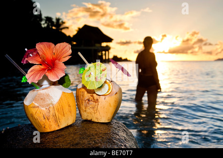 Zwei Kokosnüsse mit Cocktails und Dekorationen auf einem Granitfelsen bei Sonnenuntergang, in den Rücken, die, den eine Frau in das Meer, Seychellen steht Stockfoto