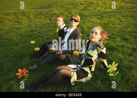 Drei Frauen sitzen nebeneinander im Feld mit Blumen, Blick in die Kamera, mit Laptop-Computer Stockfoto