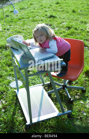 Kleines Mädchen stand auf Bürostuhl Schreibtisch im Feld, mit Laptop-Computer zu spielen, Blick in die Kamera Stockfoto