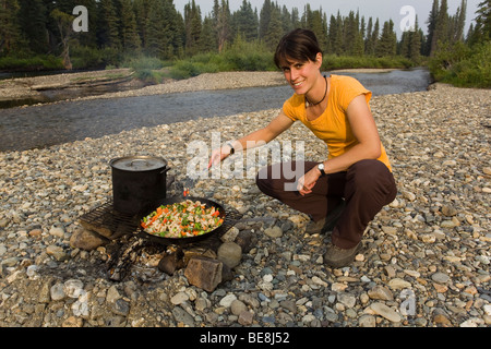 Junge Frau, Kochen, Braten Huhn rühren braten auf ein Lagerfeuer, Topf, Pfanne, Pfannenwender, Kiesbank, oben Liard River, Yukon-Territorium, Stockfoto