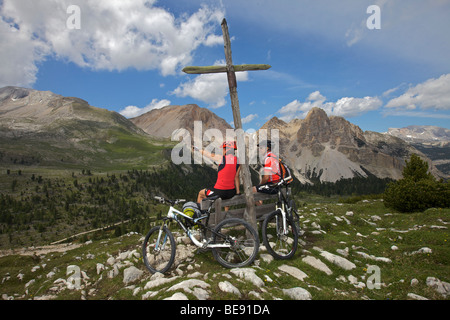 Mountainbike-Fahrer, die auf einer Holzbank vor einem Holzkreuz auf dem Limo-Pass in Fanes-Sennes-Prags Nat eine Rast machen Stockfoto