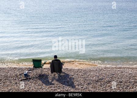 einzelne Fischer sitzen auf Stuhl am Rand des Wassers am Kiesstrand in West Bay Stockfoto