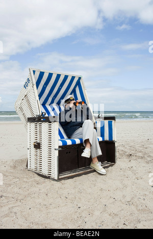 Ein senior Mann sitzt im Blick durch ein Fernglas mit Kapuze Strandkorb Stockfoto