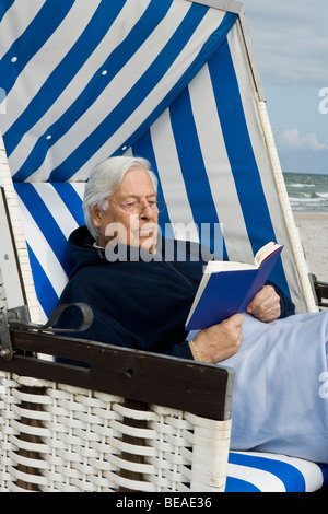 Ein senior Mann sitzt auf einem Strandstuhl mit Kapuze lesen Stockfoto