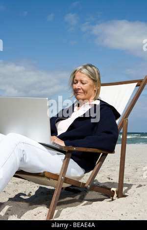 Eine ältere Frau sitzt in einem Liegestuhl am Strand mit einem laptop Stockfoto