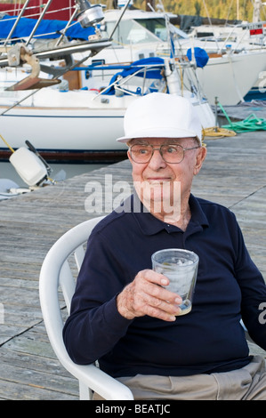 Ein gut aussehender älterer Mann hält einen Drink beim Sitzen auf einem Stuhl auf einer Marina Dock im pazifischen Nordwesten. Stockfoto