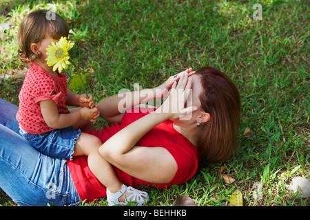 Junge Frau auf dem Rasen liegen und bedeckte ihr Gesicht mit ihren Händen und ihrer Tochter sitzt auf dem Bauch Stockfoto