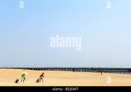 Zwei Frauen gehen auf dem Sand Strand von Coney Island in Brooklyn, New York. Eine Dame sucht Schatten unter einem Regenschirm. Stockfoto