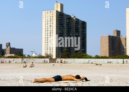 Ein Mann schläft auf den Sand am Strand von Coney Island, Brooklyn, New York. Hochhaus-Wohnprojekte sind im Hintergrund abgebildet. Stockfoto