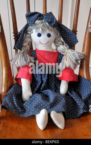 Eine hausgemachte Stoff Puppe gekleidet in blaue und rote Schürze mit einer Schleife im Haar, sitzt auf einem Holzstuhl. Stockfoto