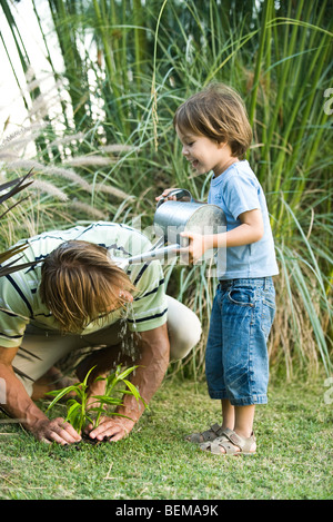 Kleiner Junge gießt Wasser auf seines Vaters Kopf, wie er im Garten arbeitet Stockfoto