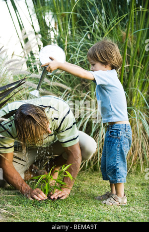 Kleiner Junge gießt Wasser auf seines Vaters Kopf, wie er im Garten arbeitet Stockfoto