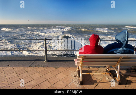 Paar Touristen auf Bank am Deich mit Blick auf die Wellen brechen über Wellenbrecher im Winter entlang der Nordseeküste Stockfoto