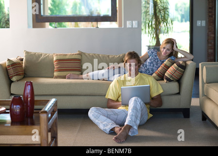 Paar zusammen im Wohnzimmer, Frau liegend auf Sofa, man sitzt am Boden mit Laptop-computer Stockfoto