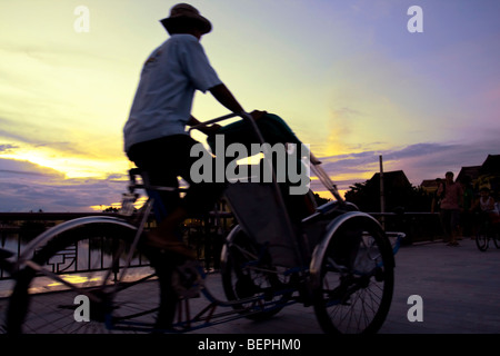 Straßenszene. Cyclo-Fahrer bei Sonnenuntergang auf einer Brücke auf dem Fluß in Hoi An. Vietnam, Südostasien Stockfoto